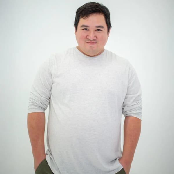 A smiling man with dark hair and a light gray long-sleeved shirt, hands in pockets.