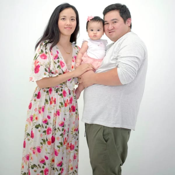 A family of three, a man, woman, and baby, pose together against a white background.