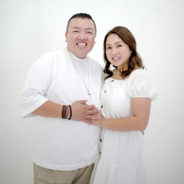 Smiling couple, a man and a woman, in white shirts, holding hands against a white background.