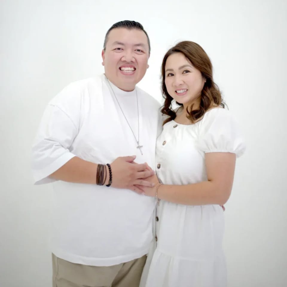 Smiling couple, a man and a woman, in white shirts, holding hands against a white background.