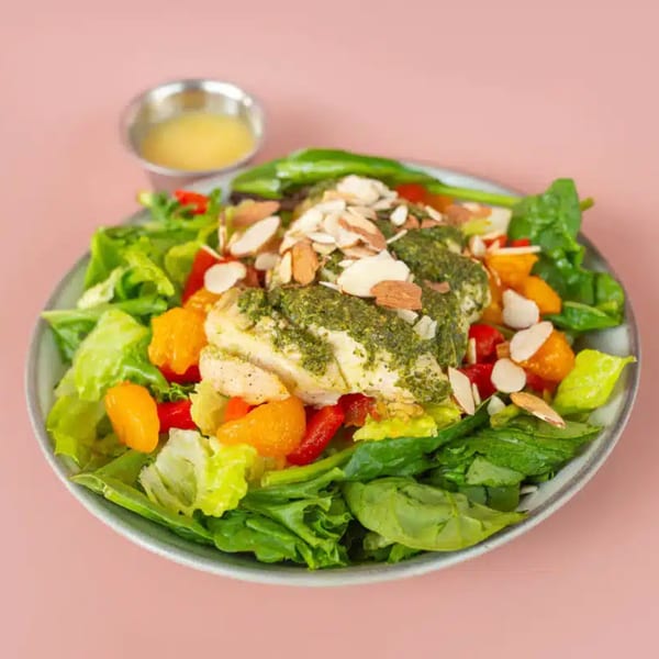 A vibrant salad with chicken, pesto, almonds, and citrus, served with dressing on a pink background.