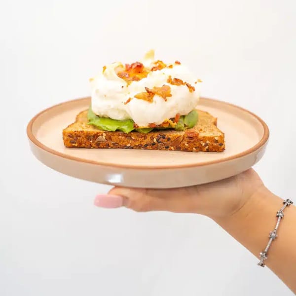 A hand holds a plate with avocado toast topped with whipped cream and bacon bits.