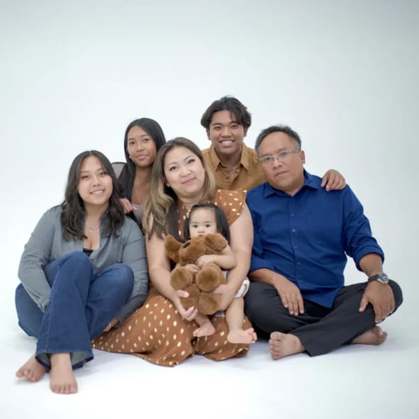 A family of six, including a baby holding a teddy bear, sits on a white floor.