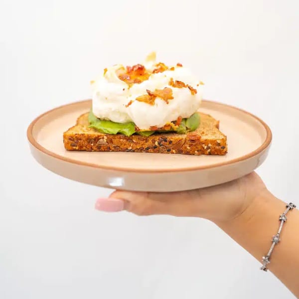 A hand holds a plate with avocado toast topped with fluffy eggs and bacon bits.