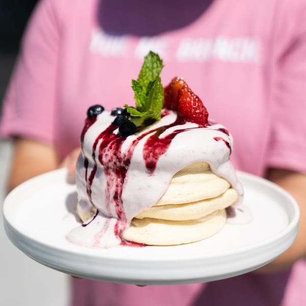 Fluffy pancakes topped with berry cream, fruit, and mint, held by a person in a pink shirt.