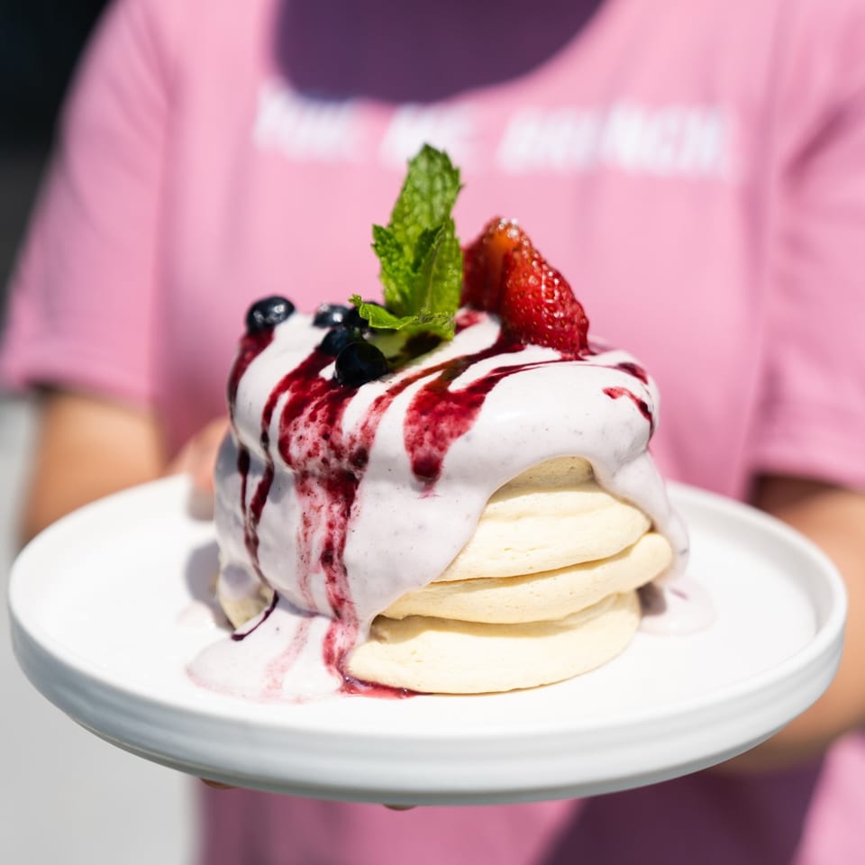 Fluffy pancakes topped with berry cream, fruit, and mint, held by a person in a pink shirt.