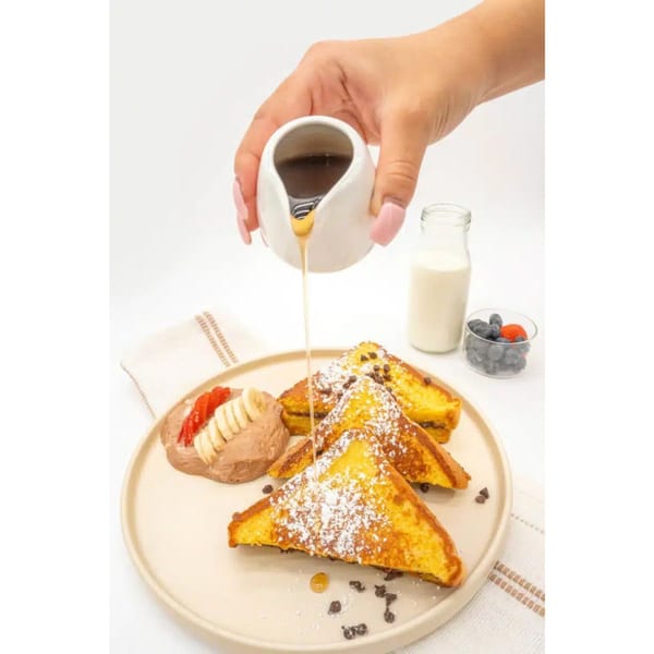 Hand pouring syrup over French toast with powdered sugar, chocolate chips, and a side of fruit.