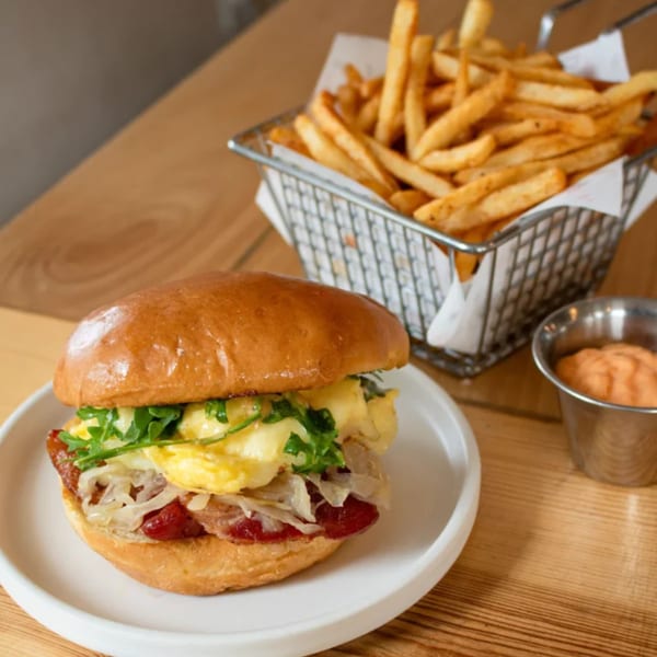 A brunch sandwich with egg, bacon, and arugula, served with a side of fries and dipping sauce.