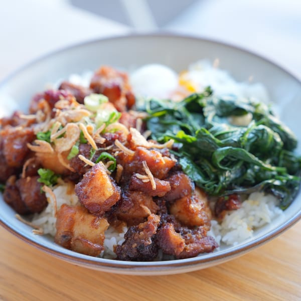 A close-up of a brunch bowl with crispy pork belly, rice, green vegetables, and a fried egg.