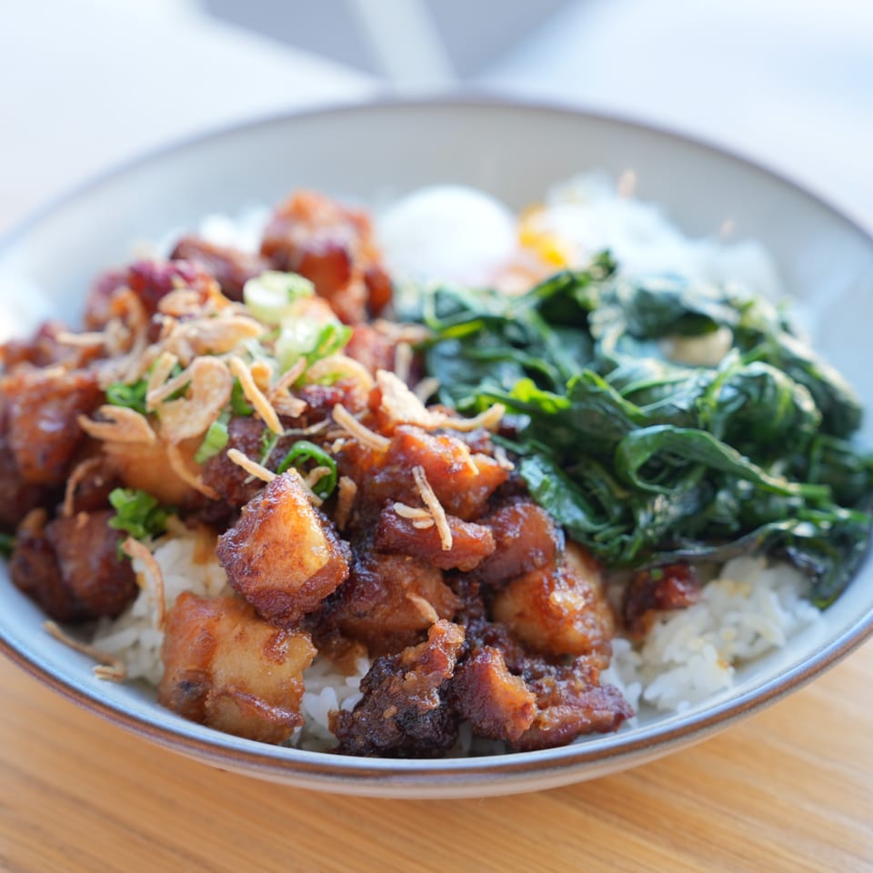 A close-up of a brunch bowl with crispy pork belly, rice, green vegetables, and a fried egg.