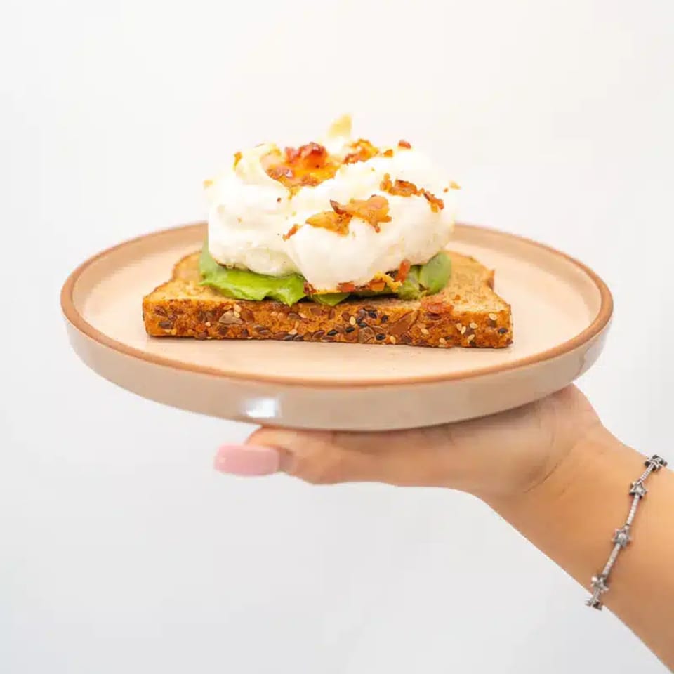 A hand holds a plate with avocado toast topped with poached eggs and bacon bits.