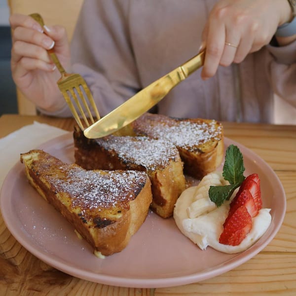 A person cuts into thick slices of French toast dusted with powdered sugar, served with fruit and cream.